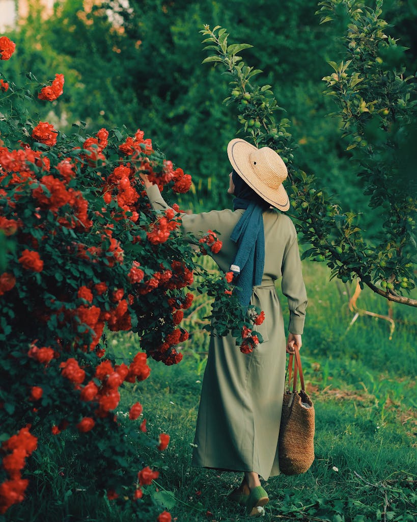 A woman in a green dress and straw hat explores a lush garden with vibrant red roses, capturing summer's beauty.
