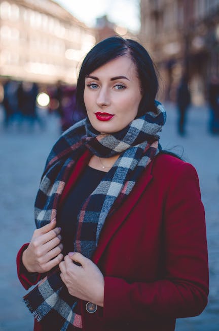 Young woman in red coat and plaid scarf standing in a bustling city street.